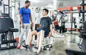 Canva: Um personal trainer e seu aluno no adutor e máquina adutora