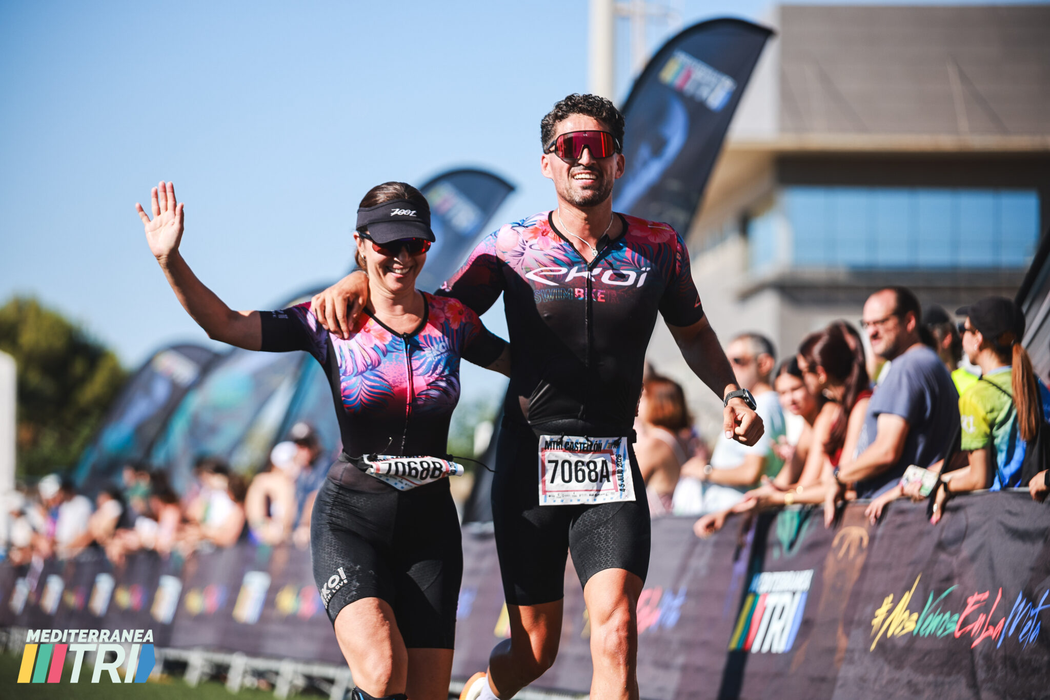 Dos participantes en una prueba de Mediterránea Triatlón avanzan juntos; una triatleta saluda mientras el público observa el recorrido.