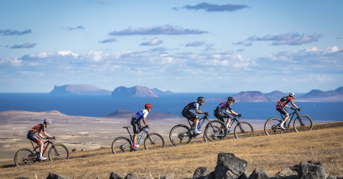 Cinco ciclistas de montaña suben por una pista de Lanzarote con el océano y el paisaje volcánico al fondo