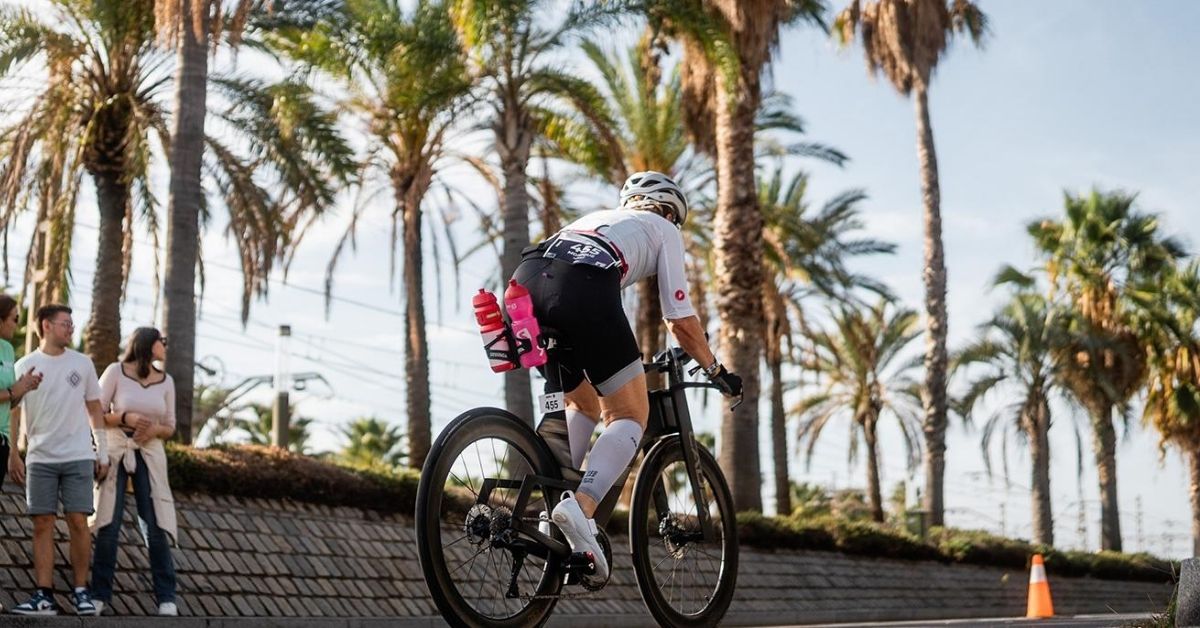 Triatleta pedaleando en una bicicleta de contrarreloj junto a un paseo con palmeras durante una competición