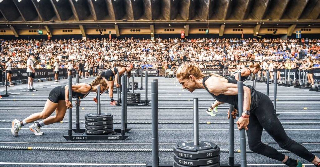 Participantes de HYROX empujan trineos con peso durante una competición de fitness híbrido en un pabellón lleno de público