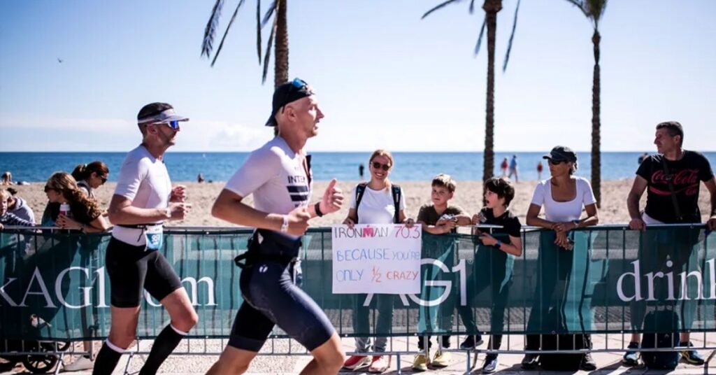 Dos triatletas corren junto a la playa durante un IRONMAN 70.3, animados por espectadores tras las vallas